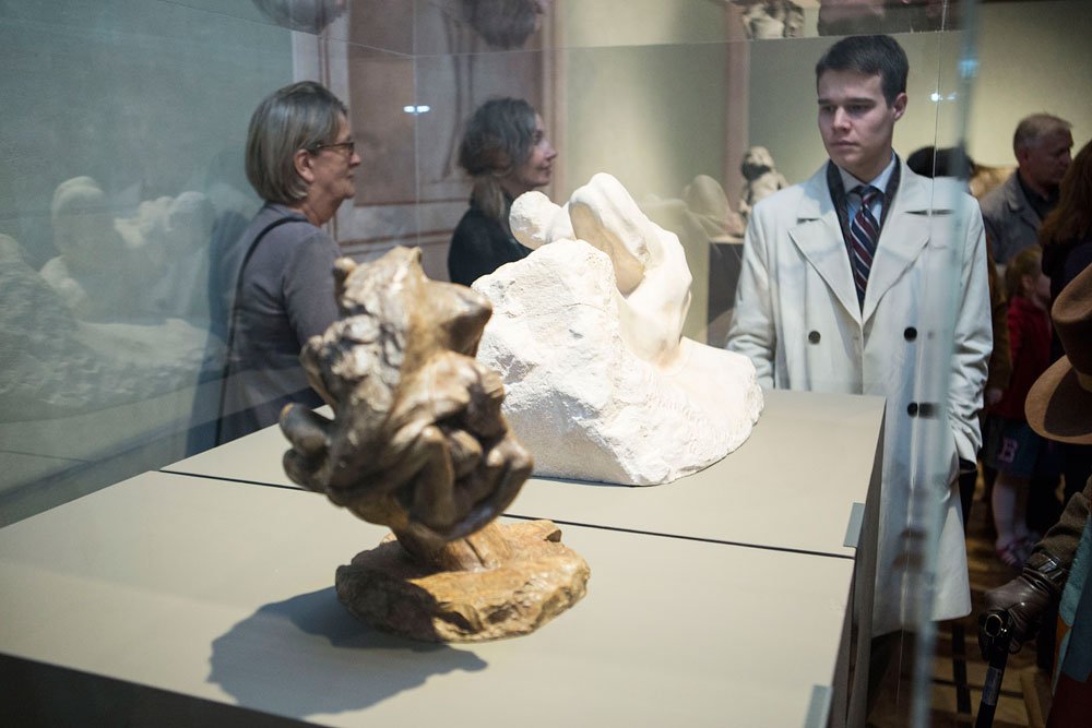 Wnętrze muzeum z dwiema rzeźbami na szarej podstawie za szklaną witryną — ciemnobrązowa, nieregularna forma na pierwszym planie i biała, gładka rzeźba za nią — oraz kilkoma zwiedzającymi w tle, w tym młody mężczyzna w jasnym płaszczu i krawacie po prawej stronie.