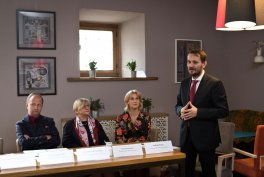 Wnętrze sali z czterema osobami przy stole — trzy osoby siedzą po lewej za stołem z tabliczkami i stosami białych dokumentów, a mężczyzna w ciemnym garniturze i krawacie stoi po prawej; w tle okno z doniczkami, ramki ze zdjęciami na ścianie i wisząca lampa.