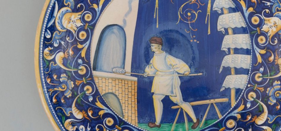 Okrągły, ozdobny ceramiczny talerz z centralnym malunkiem mężczyzny w białym stroju i brązowej czapce trzymającego długą łopatę z kilkoma bochenkami przed sklepionym otworem pieca na ciemnoniebieskim tle oraz bogato zdobioną granatowo-żółtą bordiurą z motywami roślinnymi i stylizowanymi twarzami.