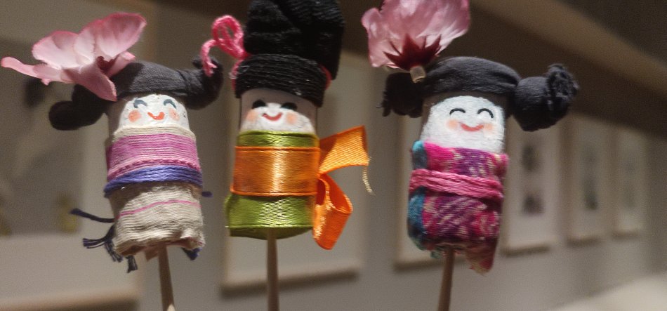 Trzy miniaturowe, ręcznie wykonane lalki na patyczkach w pierwszym planie: o białych, namalowanych twarzach z uśmiechami, owinięte kolorowymi tkaninami i wstążkami, z czarnymi włosami z nici ułożonymi w koki i różowymi papierowymi kwiatami, na rozmytym tle ściany z oprawionymi obrazami.