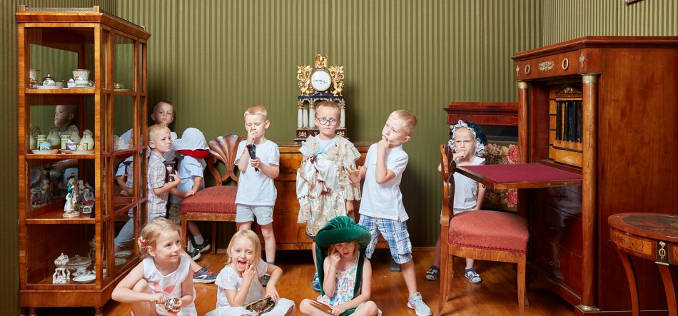 Grupa dzieci w różnym wieku ustawiona w zabytkowym pokoju z zieloną, pionowo prążkowaną tapetą i drewnianymi meblami: po lewej szklana witryna z porcelaną, pośrodku komoda z ozdobnym, złoconym zegarem, po prawej sekretarzyk z rozłożonym blatem, kilka dzieci siedzi na drewnianej podłodze z przodu, inne stoją przy meblach, niektóre mają okulary, duży zielony kapelusz lub koronkowy czepek.
