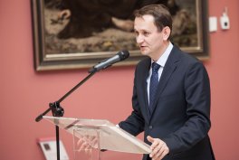 Mężczyzna w ciemnym garniturze i krawacie stoi przy przezroczystym mównicy i mówi do mikrofonu, na tle obrazu zawieszonego na różowej ścianie.