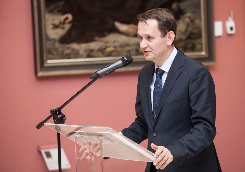 Mężczyzna w granatowym garniturze i krawacie w białe kropki stoi za przezroczystą mównicą i przemawia do mikrofonu na statywie, w tle widać oprawiony obraz na różowej ścianie.