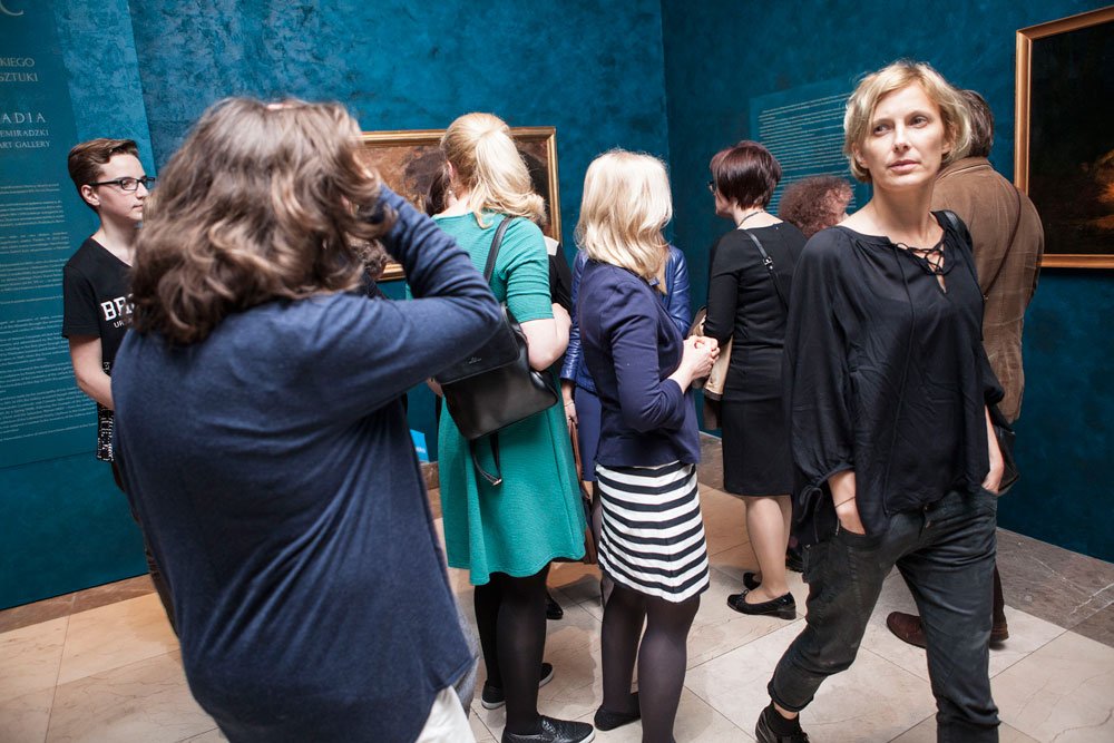 Grupa osób stojąca w sali galerii z turkusowymi ścianami i oprawionymi obrazami; z przodu po prawej kobieta w czarnej bluzce z rękami w kieszeniach, po lewej osoba od tyłu poprawiająca włosy, a w środku kilka osób w różnych strojach (zielona sukienka, granatowa marynarka, pasiasta spódnica).