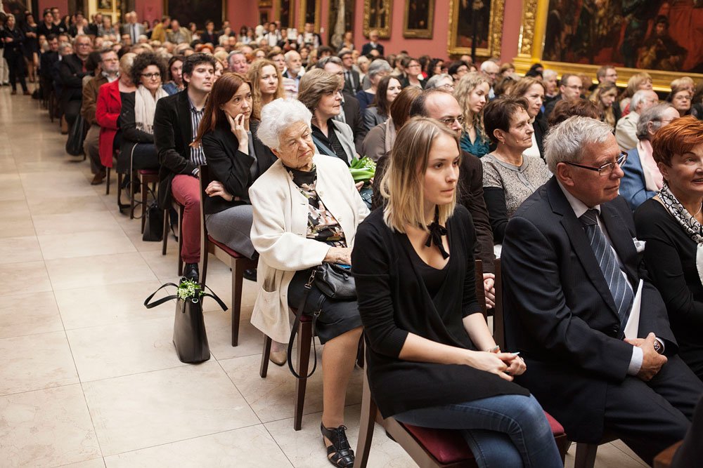 Grupa osób siedząca w wielu rzędach krzeseł w przestronnej sali z jasną posadzką i bogato zdobionymi, złoconymi ramami obrazów na ścianach; na pierwszym planie starsza kobieta w białym płaszczu, młoda kobieta w czarnej bluzce i mężczyzna w garniturze, obok na podłodze czarna torebka z wystającymi zielonymi liśćmi.