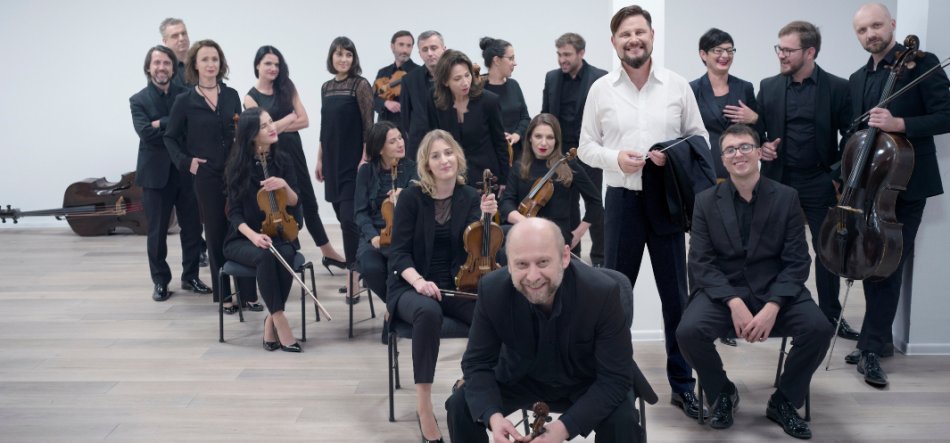 Grupa muzyków w przeważnie czarnych strojach w jasnym pomieszczeniu z jasną drewnianą podłogą i białą ścianą, część osób siedzi, część stoi, wielu trzyma instrumenty smyczkowe (skrzypce, altówki, wiolonczele, kontrabas), na pierwszym planie kucający mężczyzna trzyma skrzypce, obok stoi mężczyzna w białej koszuli trzymający marynarkę.