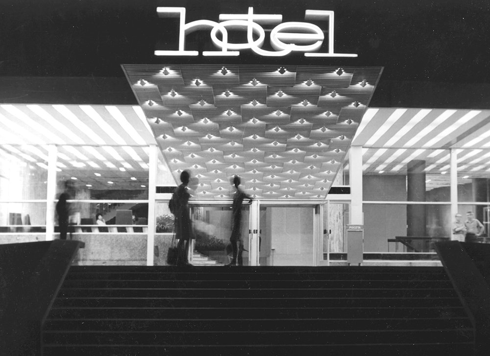Nocne wejście do hotelu z podświetlonym napisem hotel, szerokimi schodami prowadzącymi do przeszklonego lobby, geometrycznym daszkiem z rzędami punktowych świateł i kilkoma rozmytymi sylwetkami osób przy wejściu.