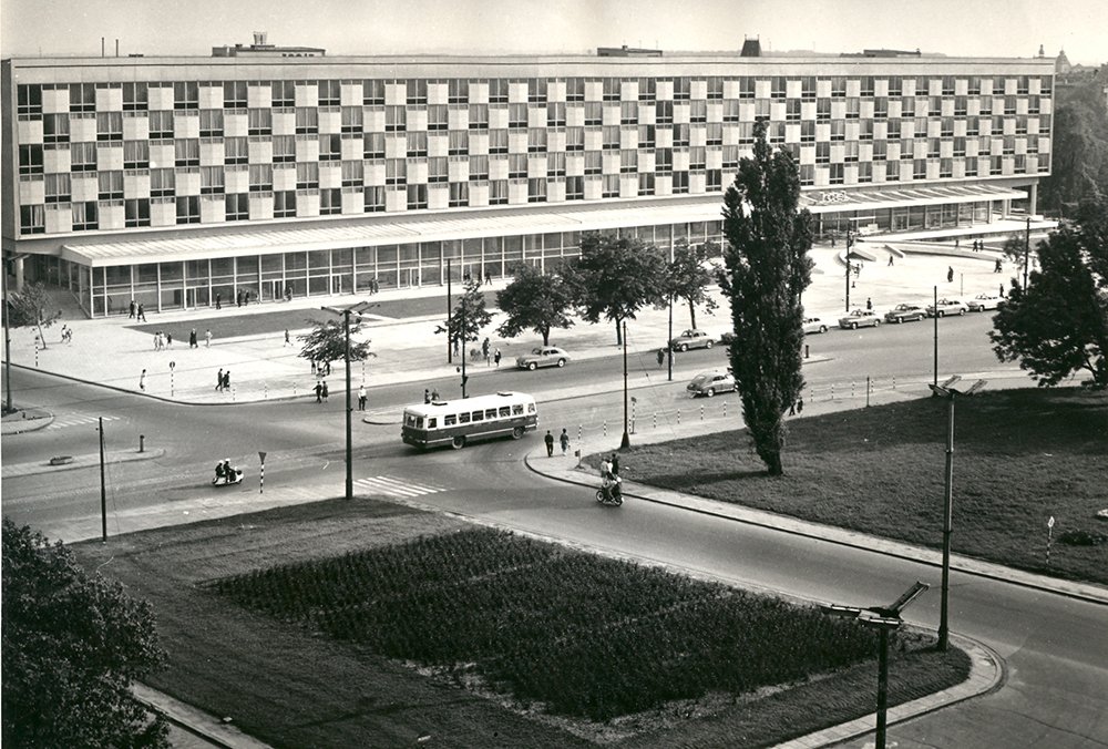 Czarno-białe zdjęcie szerokiego, wielopiętrowego budynku o regularnej, „szachownicowej” fasadzie i przeszklonym parterze, przed którym rozciąga się plac i ulica z autobusem, zaparkowanymi samochodami, przechodniami, drzewami i trawnikami.