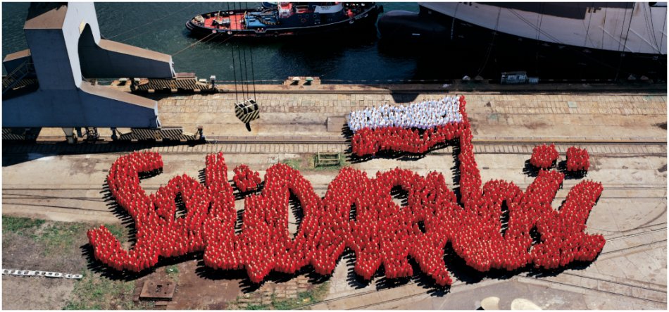 Widok z lotu ptaka na grupę ludzi ubranych na czerwono ustawionych na nabrzeżu portowym w kształt kaligraficznego napisu Solidarność, obok którego znajduje się uformowany z osób biało-czerwony kształt flagi, a w tle tory kolejowe, dźwig i cumujące statki.
