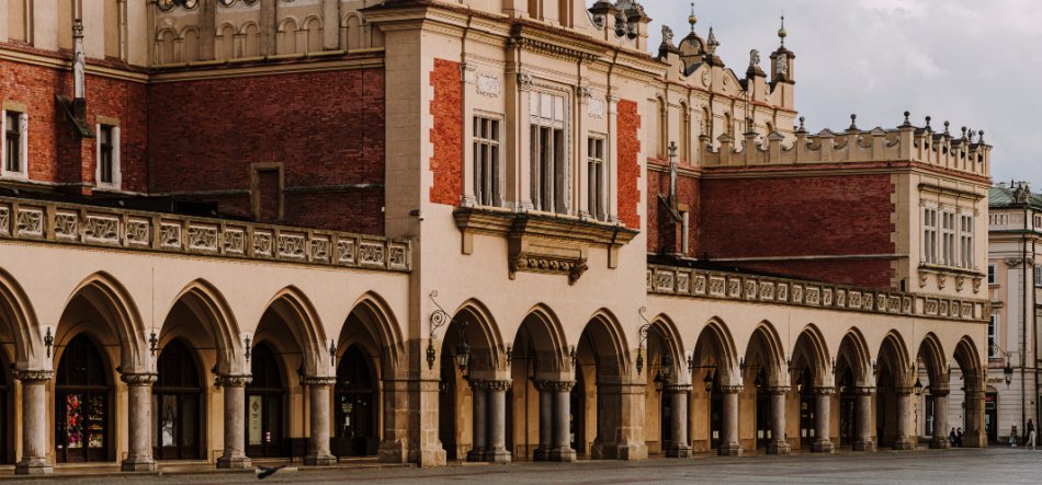 Długi zabytkowy budynek z rzędem kamiennych arkad i kolumn na parterze, ozdobną elewacją z czerwonej cegły i jasnego tynku na piętrze, metalowymi lampami przy przejściach oraz pustym brukowanym placem na pierwszym planie pod zachmurzonym niebem.
