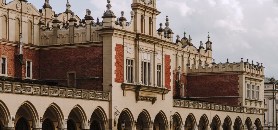 Fasada zabytkowego budynku z rzędem ostrołukowych arkad u podstawy, ceglanymi i jasnymi kamiennymi fragmentami elewacji, zdobionymi gzymsami i balustradami, ozdobnymi oknami i wieżyczkowymi sterczynami pod zachmurzonym niebem.