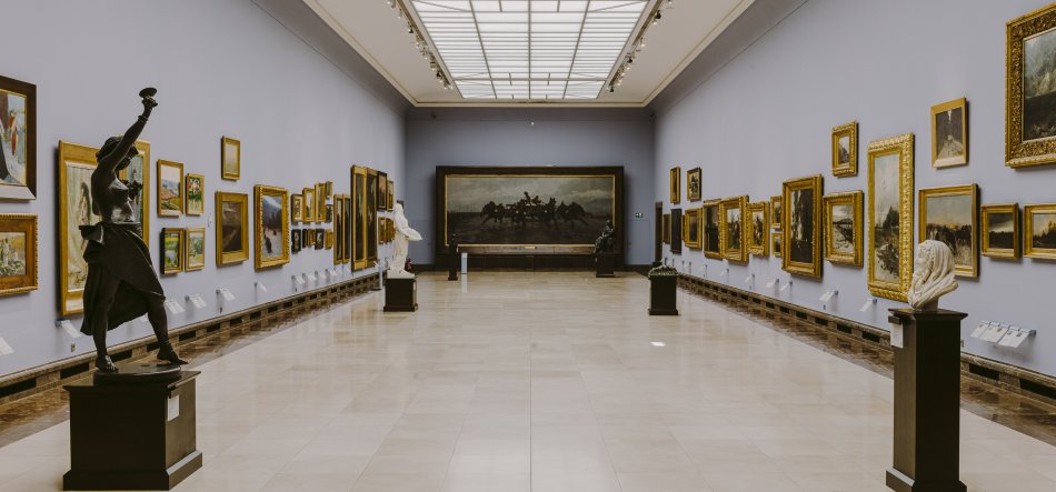 Przestronna sala galerii z dużym świetlikiem sufitowym, błękitnoszarymi ścianami i polerowaną jasną posadzką, z licznymi złoconymi obrazami w ramach po obu stronach oraz kilkoma rzeźbami na ciemnych postumentach wzdłuż środka, a na końcu wielki ciemny obraz przedstawiający jeźdźców na koniach.