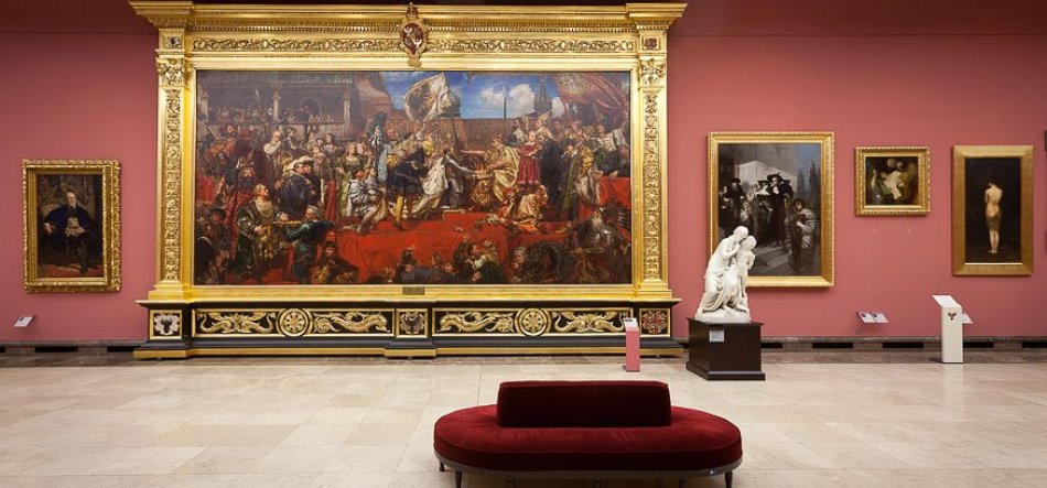 Duża sala galerii z różowawo-czerwoną ścianą; centralnie monumentalne, bogato zdobione złote ramy z dużym obrazem przedstawiającym scenę z wieloma postaciami, po prawej biała marmurowa rzeźba na cokole i kilka mniejszych obrazów w złotych ramach, po lewej kolejne malowidła, a na pierwszym planie bordowa owalna kanapa na jasnej kamiennej posadzce i niewielkie tabliczki informacyjne przy ścianach.