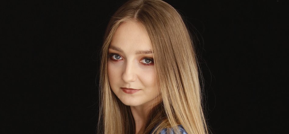 Zbliżony portret młodej kobiety na czarnym tle, z długimi prostymi blond włosami, jasną cerą, subtelnym makijażem z ciemniejszym cieniem na powiekach, lekko odwróconą twarzą w stronę aparatu i ubraniem w niebieskim kolorze.