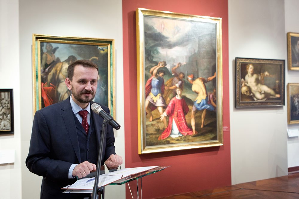 Mężczyzna w ciemnym garniturze i bordowym krawacie stoi przy mikrofonie i pulpicie z kartkami na tle galerii z kilkoma oprawionymi, klasycznymi obrazami, w tym dużym obrazem figuralnym na czerwonej ścianie.