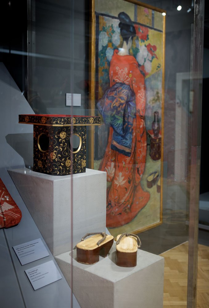 Ekspozycja w muzeum za szybą: na białych postumentach ustawione drewniane sandały geta z podwyższonymi podeszwami i sznurkami oraz czarno-złota szkatułka z ornamentami, a w tle oprawiony obraz przedstawiający kobietę w czerwonym kimonie; przy eksponatach widoczne tabliczki informacyjne i odbicia w szkle.