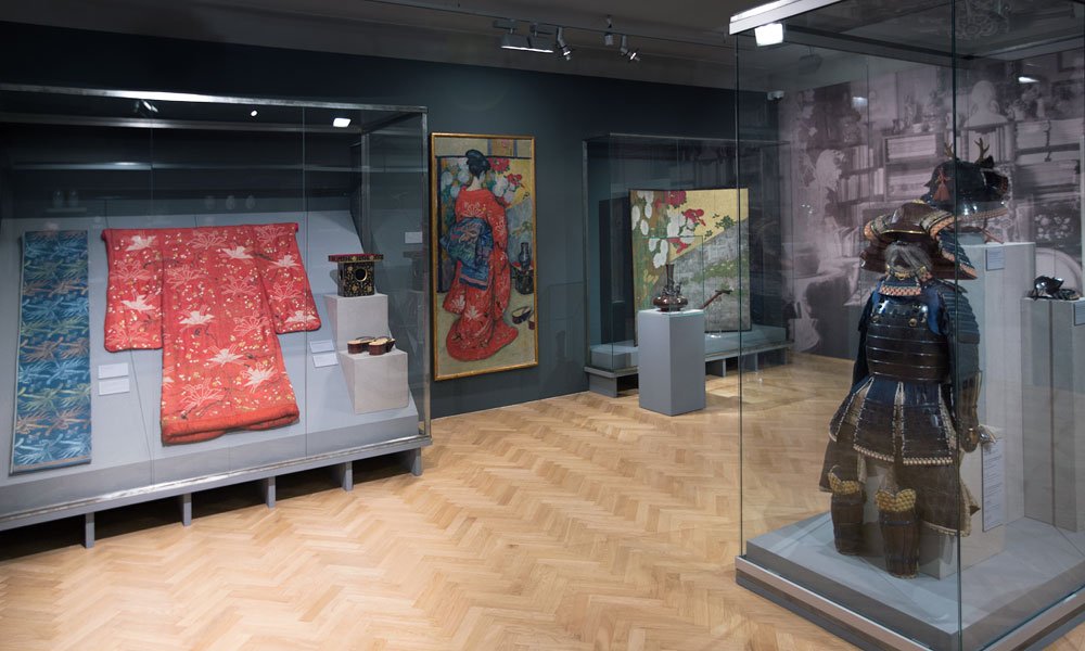 Sala wystawowa z drewnianą jodełkową podłogą i przeszklonymi gablotami, w których po lewej eksponowane jest czerwone kimono i tkaniny, na ścianie wisi obraz, a po prawej w osobnej gablocie stoi tradycyjna zbroja z hełmem.