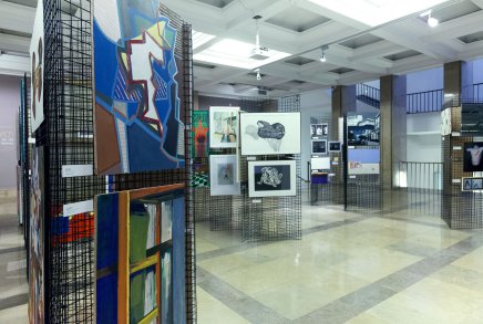 Wnętrze jasnej galerii wystawowej z czarnymi metalowymi panelami kratowymi ustawionymi w przestrzeni sali, na których wiszą kolorowe obrazy i oprawione czarno-białe grafiki, widoczne jasne płytki podłogowe, sufit z kwadratowymi wnękami i punktowymi lampami oraz barierka i okna na półpiętrze w tle.