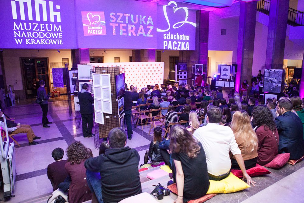 Wnętrze sali Muzeum Narodowego w Krakowie podczas wydarzenia: publiczność siedzi na krzesłach i poduszkach na stopniach przed sceną i ekspozycjami na metalowych stojakach, a na ścianie widoczne są fioletowe projekcje z napisami "Muzeum Narodowe w Krakowie", "Sztuka Teraz" i "Szlachetna Paczka".