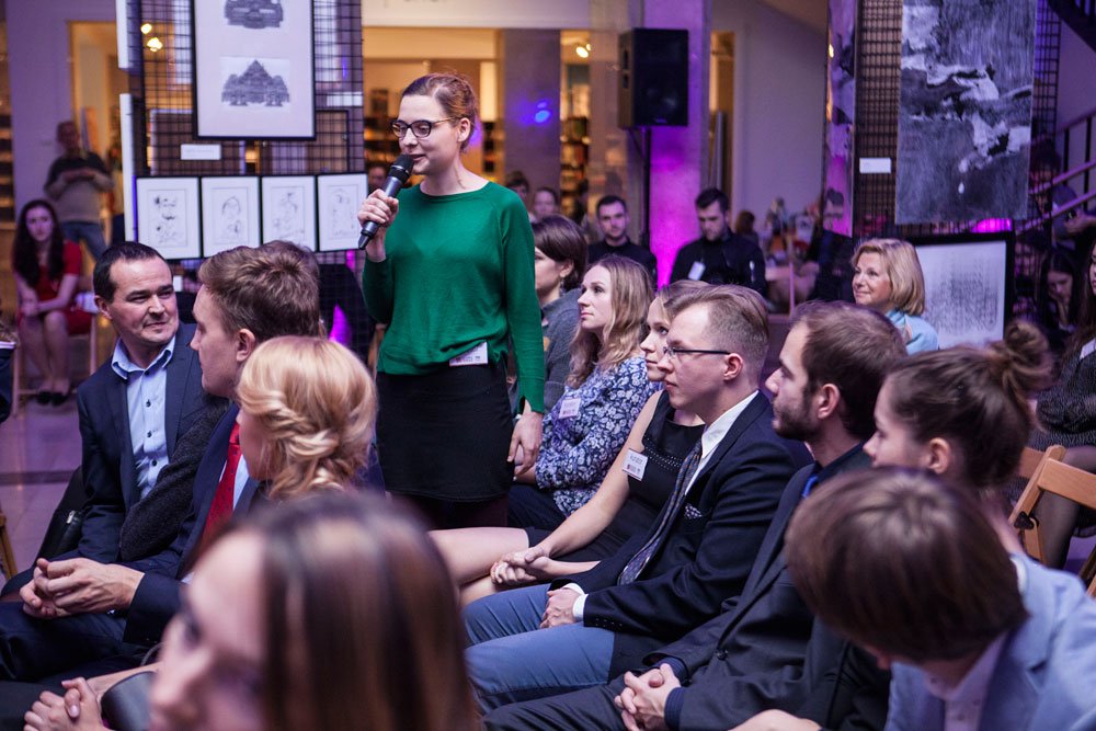 Kobieta w zielonym swetrze i okularach stoi między siedzącymi uczestnikami i trzyma mikrofon w sali z wystawą rysunków na panelach, wokół niej ludzie siedzą w rzędach, niektórzy mają identyfikatory, a wnętrze jest podświetlone fioletowym światłem.