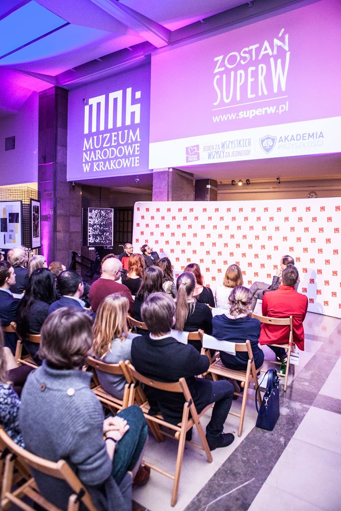 Wnętrze sali muzealnej oświetlone fioletowo, widoczne rzędy publiczności siedzącej na drewnianych składanych krzesłach skierowane ku scenie z panelem dyskusyjnym przed białym panelem z czerwonymi logotypami, a na ścianie duże projekcje z napisami Muzeum Narodowe w Krakowie i Zostań SUPERW