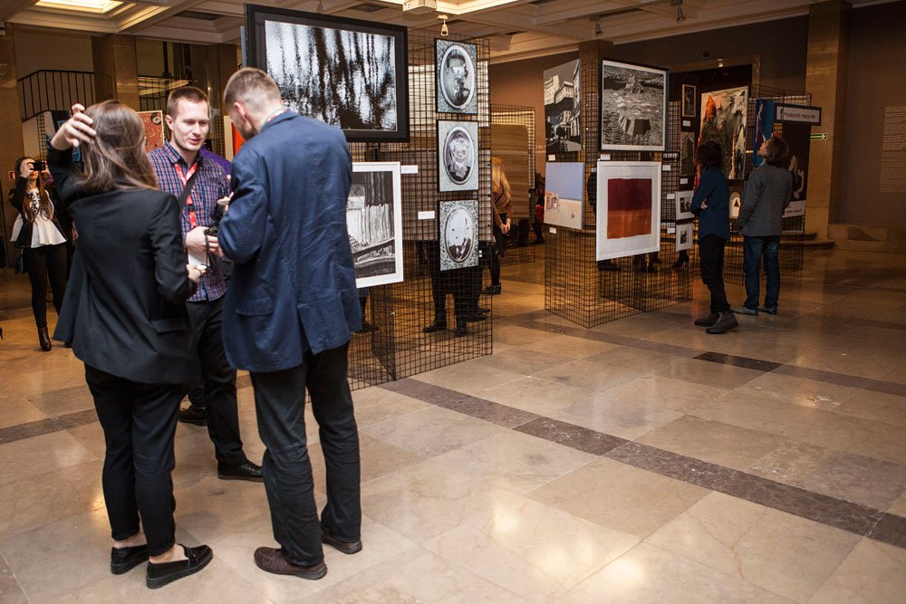 Wnętrze galerii z wystawą fotografii i grafik zawieszonych na czarnych metalowych kratach; w pierwszym planie trzy osoby rozmawiają stojąc na jasnej marmurowej posadzce, w tle inni zwiedzający oglądają prace, a jedna osoba robi zdjęcie.
