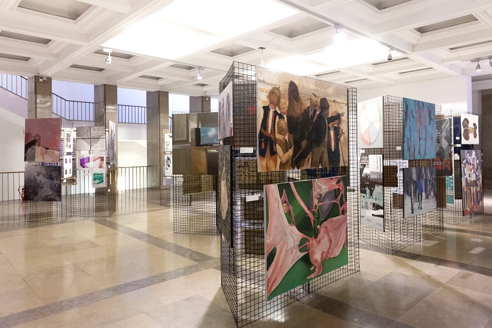 Wnętrze galerii z jasnym kasetonowym sufitem i polerowaną podłogą, z wolnostojącymi metalowymi kratowymi ściankami ustawionymi w rzędach i na nich zawieszonymi różnobarwnymi obrazami i fotografiami — w tym figuratywnym obrazem grupy postaci w strojach formalnych, dużym abstrakcyjnym płótnem w zielono‑różowych tonach oraz kilkoma innymi pracami rozmieszczonymi przy kolumnach i balustradzie.