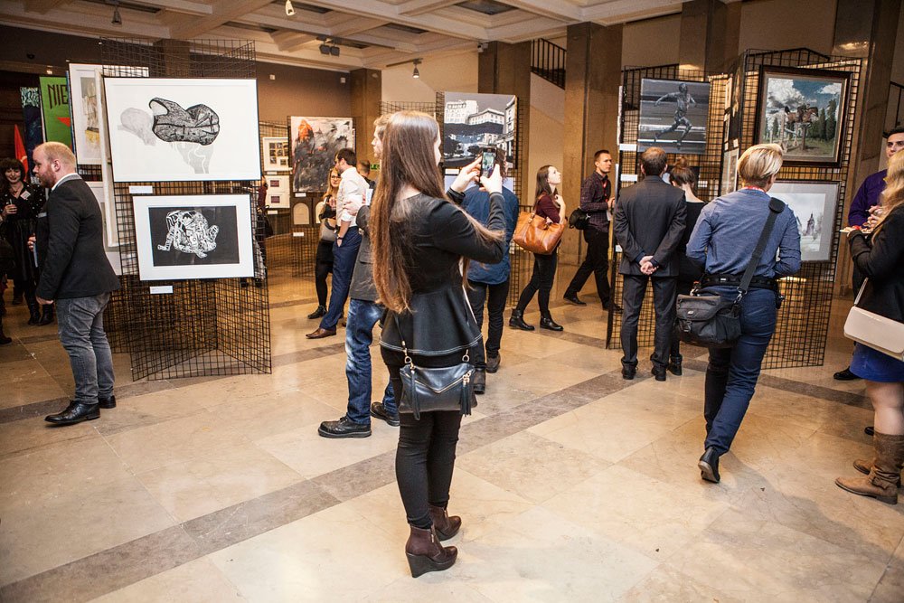 Wnętrze galerii z wystawą obrazów na metalowych panelach i kamienną posadzką, kilka osób ogląda prace — na pierwszym planie kobieta z długimi włosami w czarnym płaszczu i skórzaną torebką robi zdjęcie telefonem, w tle inni zwiedzający i oprawione grafiki oraz obrazy zawieszone na stojakach.