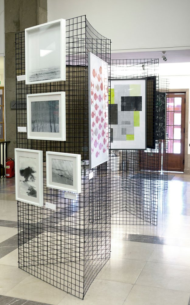 Wnętrze przestrzeni wystawowej z czarnymi siatkowanymi panelami ustawionymi w zygzak, na których zawieszone są białe ramy z czarno-białymi fotografiami oraz kolorowe grafiki (wzór różowych ryb i geometryczne bloki z zielonymi akcentami), z marmurową podłogą, drzwiami i gaśnicą w tle.