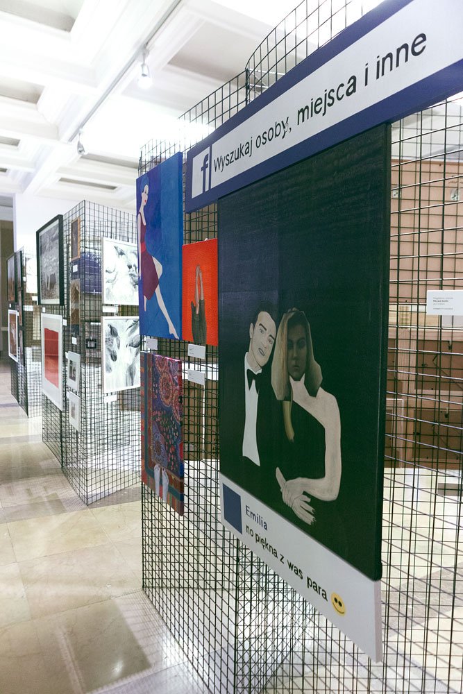 Wnętrze galerii z rzędem metalowych siatkowych paneli prezentujących obrazy; na pierwszym planie duży obraz stylizowany na wpis z Facebooka z nagłówkiem Wyszukaj osoby, miejsca i inne oraz podpisem Emilia no piękna z was para 😊 przedstawiający dwie postacie w eleganckich strojach, w tle kolejne kolorowe prace, sufit z lampami i marmurowa podłoga.