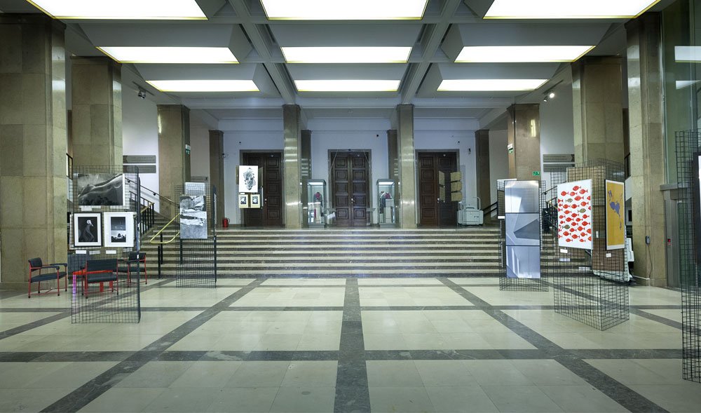 Duża jasna sala z marmurową podłogą w ciemne pasy, wysokimi kamiennymi kolumnami, siatkowymi panelami wystawowymi z fotografiami i plakatami po obu stronach, kilkoma krzesłami po lewej oraz szerokimi schodami wiodącymi do trzech dużych drewnianych drzwi na końcu i prostokątnymi świetlami wpuszczonymi w sufit.