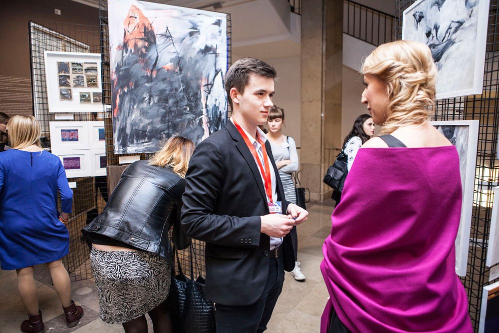 Grupa osób w galerii stoi przed metalowymi panelami z abstrakcyjnymi obrazami; na pierwszym planie mężczyzna w czarnym garniturze z czerwonym identyfikatorem rozmawia z kobietą o plecionych blond włosach owiniętą fuksjowym szalem, a wokół inni zwiedzający oglądają prace.