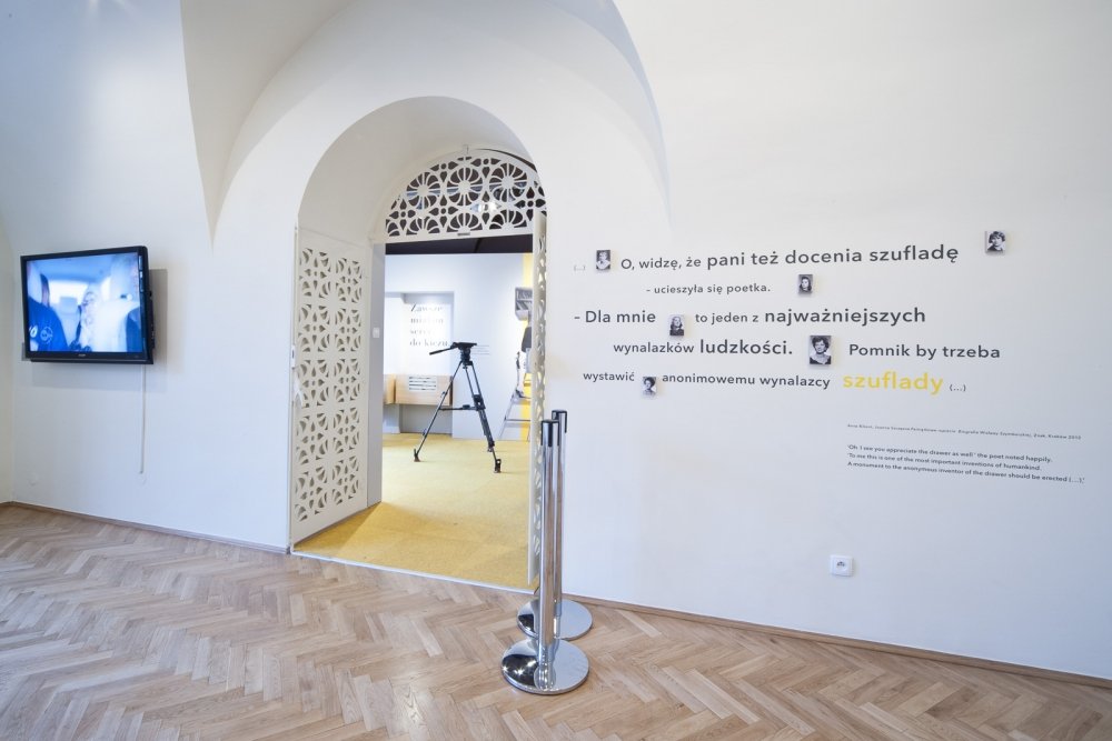 Wnętrze białej galerii z łukowym przejściem ozdobionym ażurowymi drzwiami, przez które widać żółty dywan i statyw z kamerą; po lewej wiszący ekran telewizyjny, po prawej ściana z cytatem w języku polskim i chromowanymi słupkami z taśmą, na podłodze jodełkowy parkiet.