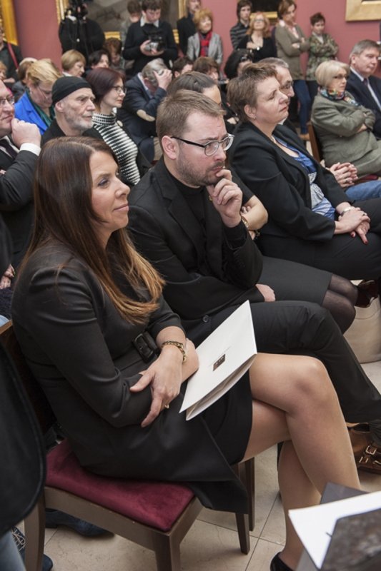 Grupa osób siedzących w rzędach w sali, na pierwszym planie kobieta w czarnej sukience z długimi brązowymi włosami trzymająca białą teczkę na udach na czerwonym krześle, obok niej mężczyzna w czarnym ubraniu i okularach z dłonią przy twarzy, w tle więcej siedzących i stojących osób, niektórzy z aparatem, oraz obrazy na ścianach.