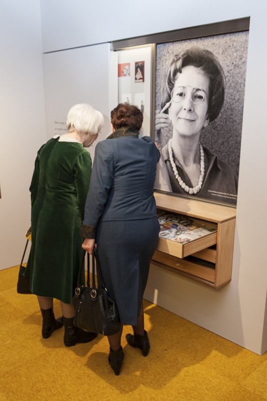 Dwie starsze kobiety widziane od tyłu, w zielonej i granatowej sukni, pochylone nad wysuniętymi drewnianymi szufladami z materiałami ekspozycyjnymi pod dużym czarno-białym portretem kobiety w okularach i koralach, stoją na żółtym dywanie.