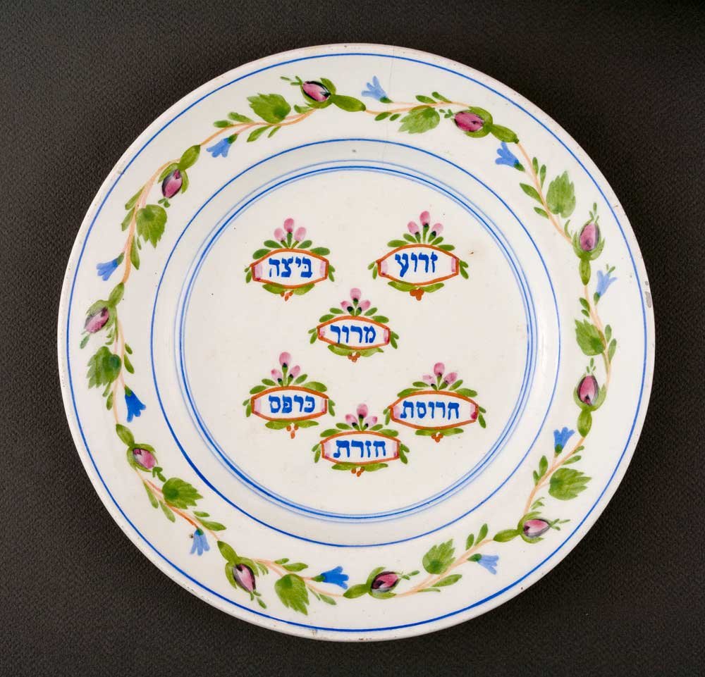 Biały okrągły ceramiczny talerz z cienkimi niebieskimi obwódkami i zewnętrznym wieńcem zielonych liści, różowych pąków i niebieskich kwiatuszków oraz siedmioma małymi ozdobnymi emblematami w środku z różowo‑zielonymi akcentami zawierającymi hebrajskie napisy.