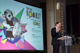 Mężczyzna stoi przy przezroczystej mównicy z mikrofonem na scenie obok dużego kolorowego ekranu projekcyjnego z komiksową ilustracją perkusisty i napisem "teraz KOMIKS".