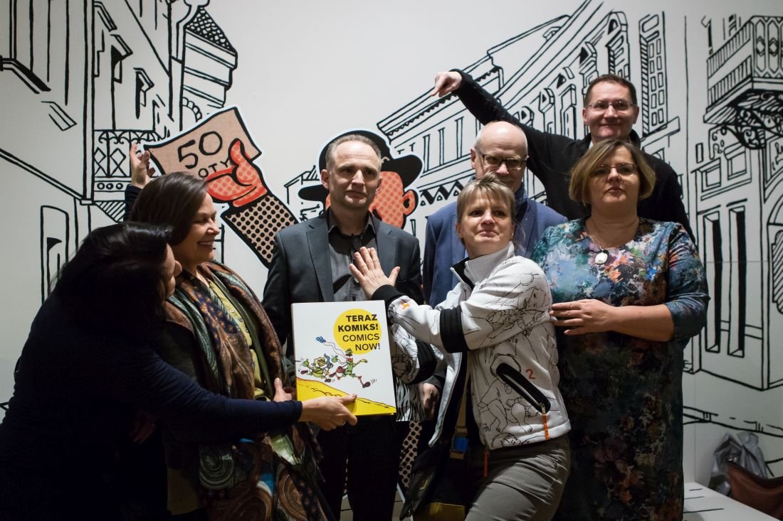 Grupowe zdjęcie siedmiu osób ustawionych na tle czarno-białej muralowej ilustracji miasta, z mężczyzną w ciemnym garniturze i kapeluszu na środku, kobietą w białej kurtce dotykającą jego klatki piersiowej i inną osobą trzymającą z przodu żółtą książkę z napisem „TERAZ KOMIKS! COMICS NOW!”, a w tle rysunek ręki trzymającej banknot z cyfrą 50 oraz zarysy budynków.