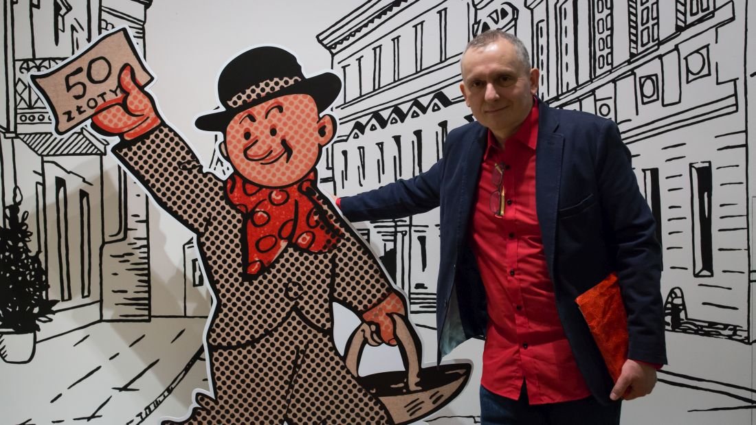 Mężczyzna w czerwonej koszuli i ciemnej marynarce stoi obok dużej kartonowej kreskówkowej postaci w kapeluszu i czerwonej chustce, która trzyma kapelusz i kartkę z napisem 50 zł, na tle czarno-białych rysunkowych kamienic.