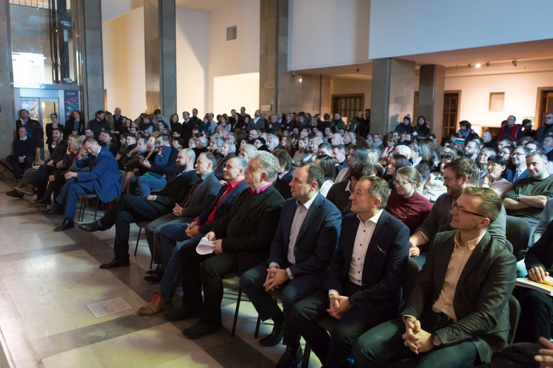 Duża grupa osób siedzących i stojących w rzędach w przestronnej, jasnej sali z wysokim sufitem i kolumnami, skierowanych w lewo; na pierwszym planie kilku mężczyzn w garniturach siedzących w pierwszym rzędzie.