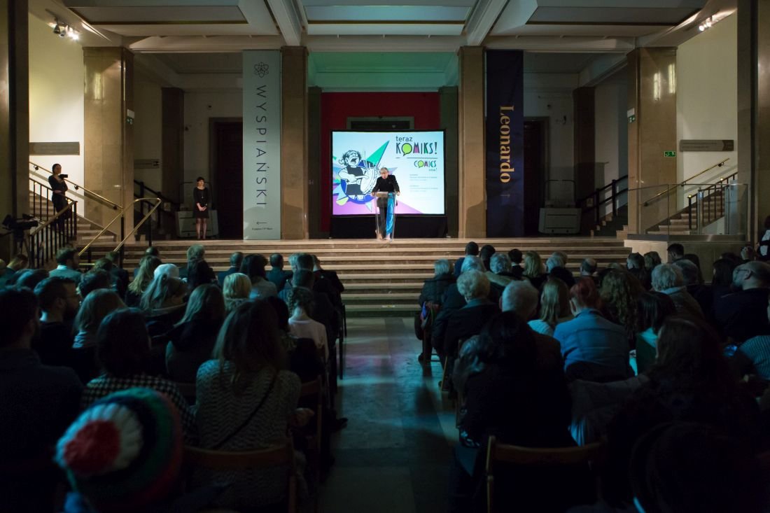 Duża sala z publicznością siedzącą w rzędach, na podwyższonej scenie osoba przy mównicy przed podświetlonym ekranem z kolorową grafiką i napisami, po bokach wysokie pionowe banery z napisami „Wyspiański” i „Leonardo” oraz schody prowadzące na scenę.