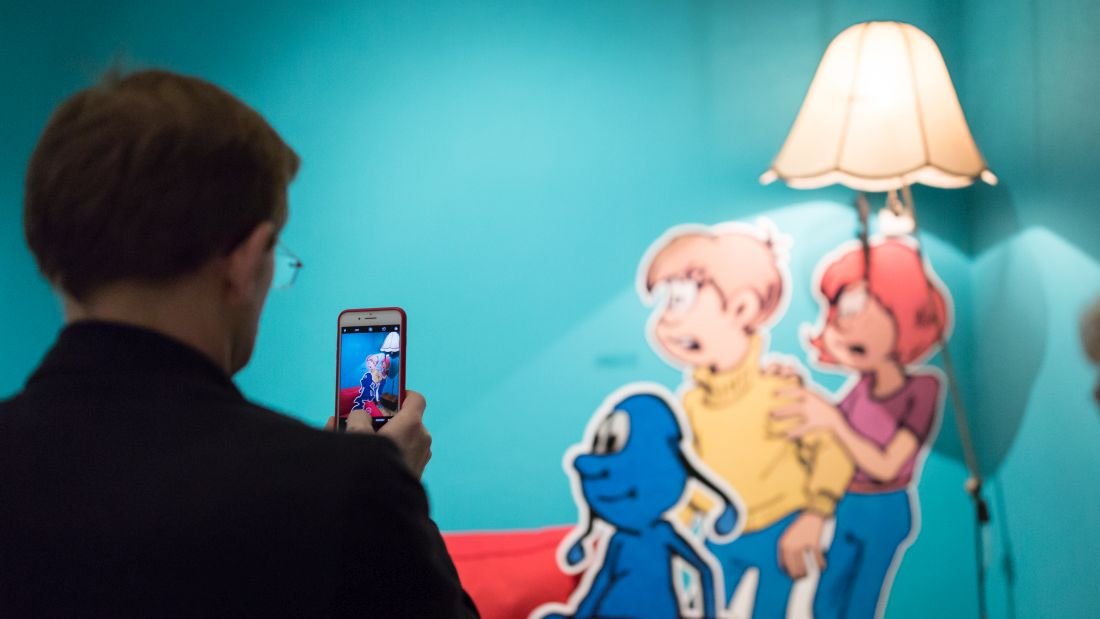 Osoba widziana od tyłu trzyma smartfon, którym fotografuje kolorowe wycięte ilustracje na turkusowej ścianie — niebieską postać i dwie postacie ludzkie (blond w żółtym swetrze i ruda w fioletowym topie) obok stojącej lampy z beżowym abażurem.