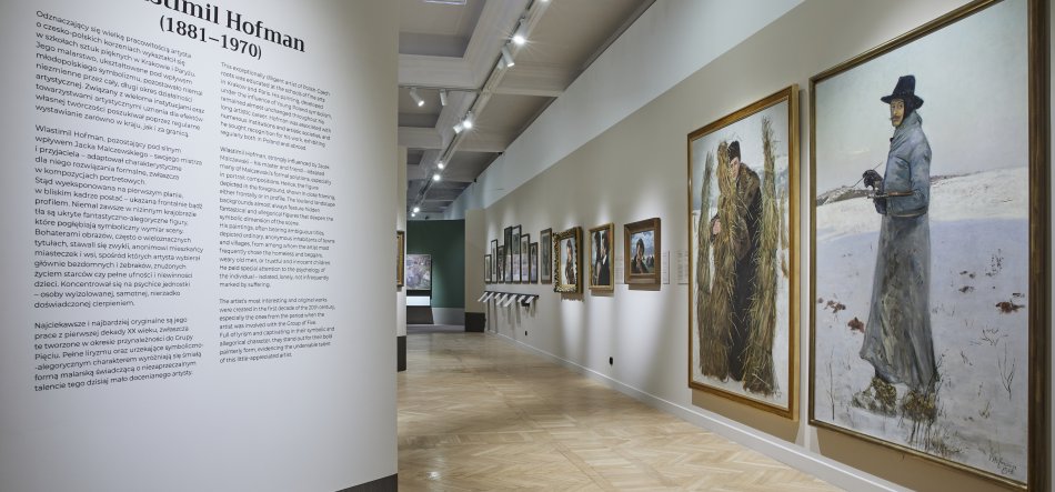 Wnętrze galerii z jasną drewnianą podłogą i sufitowymi reflektorami, po prawej ścianie rząd oprawionych obrazów widocznych w perspektywie — na pierwszym planie dwa duże płótna: jedno ukazujące postać w długim płaszczu i kapeluszu stojącą na zaśnieżonym tle i trzymającą długi przedmiot, drugie przedstawiające dwie postacie przysłonięte snopem siana — a po lewej duża informacyjna tablica z tekstem i napisem "Vlastimil Hofman (1881–1970)".
