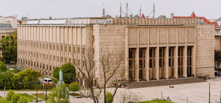 Duży budynek z jasnego kamienia o prostokątnej bryle, frontowy portyk z kilkoma wysokimi kolumnami i szerokimi schodami, długie wąskie okna po lewej stronie oraz anteny na dachu; na pierwszym planie plac z drzewami, figurą pomnikową, kilkoma zaparkowanymi samochodami i nielicznymi osobami siedzącymi na kamiennych stopniach.