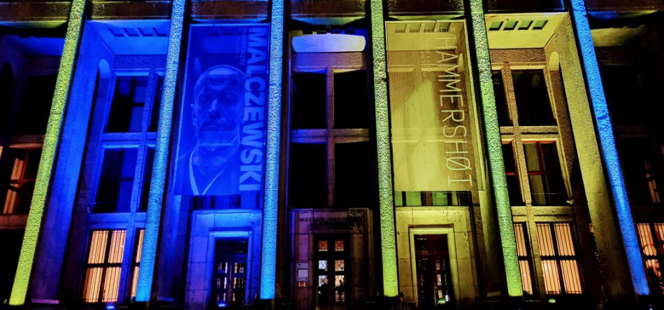 Fasada budynku z wysokimi pionowymi kolumnami oświetlonymi na niebiesko i zielono, z dwoma dużymi pionowymi banerami — na lewym widoczny podświetlony portret i napis "MALCZEWSKI", na prawym pionowy napis "HAMMERSHØI" — oraz podświetlone drzwi i okna na nocnym tle.
