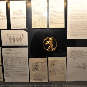 W gablocie na ciemnym tle rozmieszczone kilkanaście kartek z odręcznymi notatkami, drukowanymi tekstami i szkicami, pośrodku okrągły złotawy emblemat z wypukłą figurą zwierzęcia, na górnych arkuszach widoczny refleks świetlny, a u dołu wąska etykieta z opisem.
