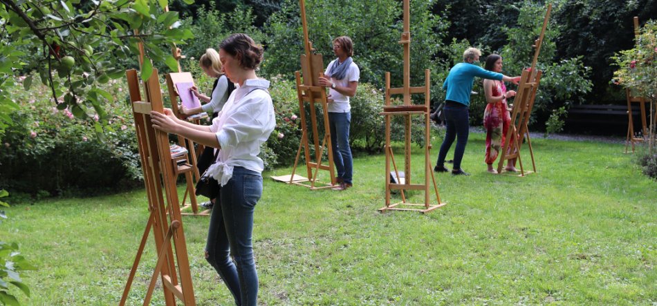 Kilka osób stoi przy drewnianych sztalugach na trawniku w ogrodzie, każda pracuje przy swoim podobraziu, w tle gęste krzewy i drzewa.