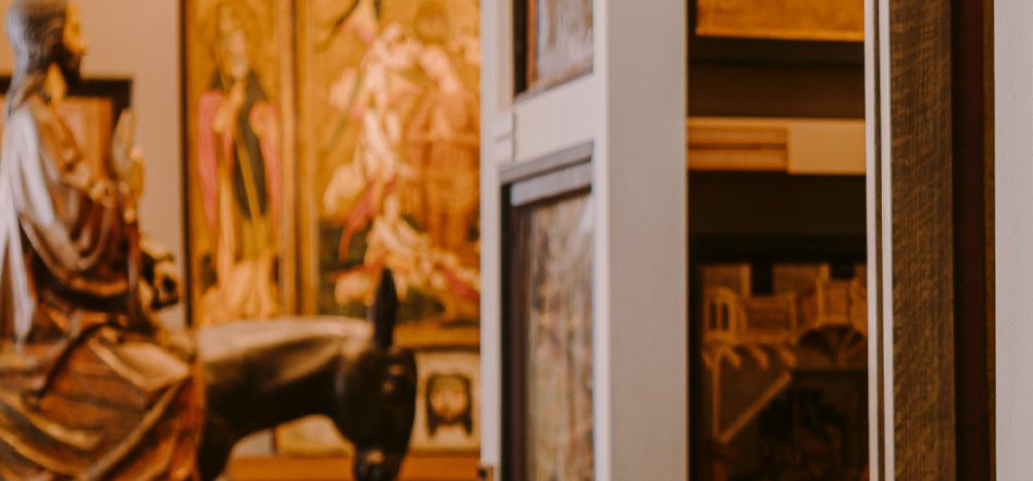 Rozmyte zdjęcie wnętrza wystawy muzealnej: po lewej rzeźba siedzącej postaci i widoczna głowa konia, w tle złociste malowidła w ramach (tryptyk), po prawej fragment przeszklonej gabloty i drewnianych ram.