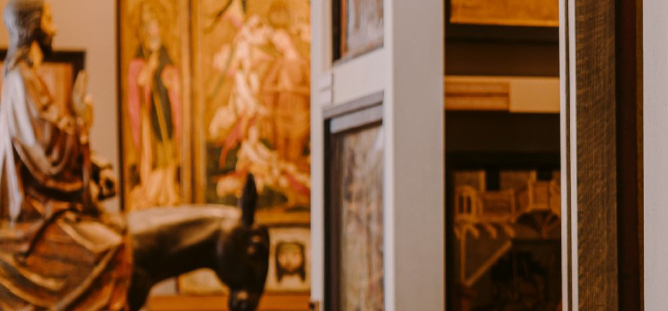 Fragment wnętrza galerii lub muzeum: w rozmytym pierwszym planie rzeźba postaci siedzącej na zwierzęciu, w tle złociste panele z przedstawieniami postaci, a po prawej wąski otwór z widocznymi oprawionymi obrazami na regale.