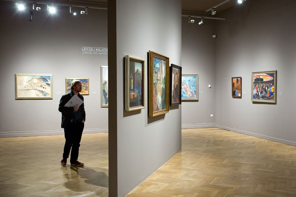 Wnętrze galerii sztuki z jasnym drewnianym parkietem, centralną wolnostojącą ścianką z kilkoma oprawionymi obrazami, dodatkowymi obrazami na tylnej i bocznej ścianie, widocznym napisem "LIRYZM I MALARST..." nad jedną ścianą oraz stojącym mężczyzną trzymającym kartki.